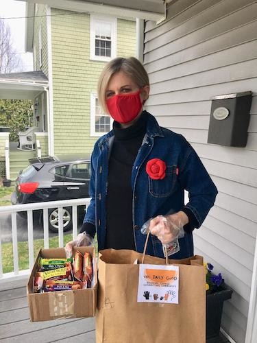 Sandra on porch wearing mask and holding a bag and cardboard box full of donations.